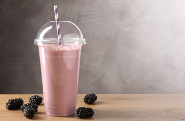 Tasty milkshake in plastic cup and blackberries on wooden table, space for text