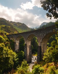 A vast, arched railway viaduct spans a valley nestled between lush, green hills under a bright, sunny sky.