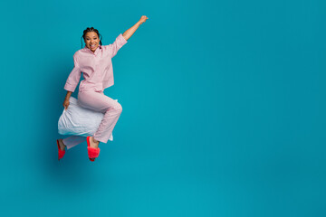 Young cheerful woman in pink pyjamas wakes up energetically on blue background while holding a...