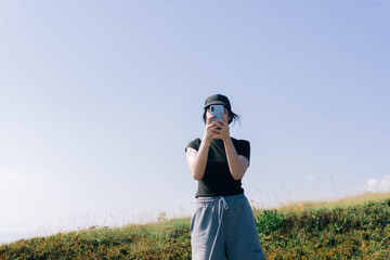 A woman stands on a hill covered in grass, holding her phone to take a selfie while wearing...