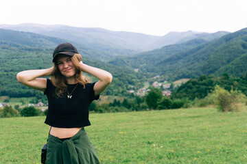A woman with long hair stands in a green field, smiling while holding her hair back. The backdrop features rolling hills and a quaint village nestled in the valley under an overcast sky.