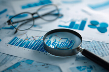 Magnifying glass over financial charts and graphs with eyeglasses in background for business analysis