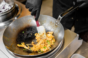 Chef stir-frying rice noodles with shrimp and herbs in a wok. Asian street food preparation with...