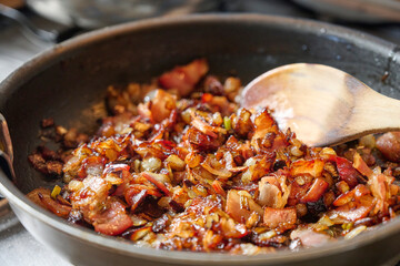 Bacon and caramelized onion cook slowly in a pan, stirred with a wooden ladle in a traditional...