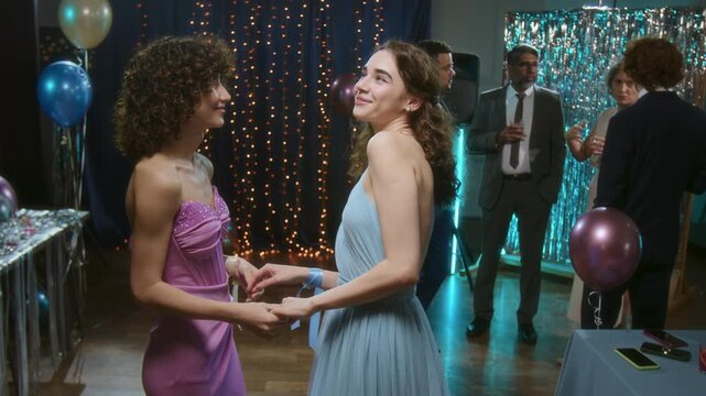 Two young female friends, wearing evening dresses, talking while playfully dancig at high school prom party
