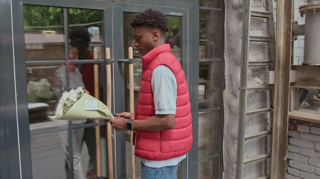 African American male courier delivering bouquet of fresh white daises to mature Caucasian woman. He knocking on door and waiting for woman
