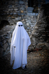 A child, draped in a flowing white sheet and wearing sunglasses, stands against a stone wall.