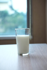 A full glass of fresh milk resting on a light wooden table, with a blurred window and wall in the background.