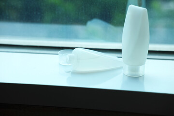 White plastic tubes of bath essentials placed near a window, one standing and one lying with open cap and liquid drop, representing shampoo, shower gel, or body lotion packaging.
