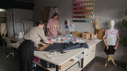 Medium full shot of young Caucasian seamstress drawing pattern on cloth with chalk in artisan workshop, constructing new dress design, while colleague is pinning together pieces of fabric