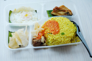 A pre-packaged meal tray containing yellow rice with sauce, a fried egg, and a piece of chicken, served on a light tabletop. Crackers and another food item are in separate compartments.