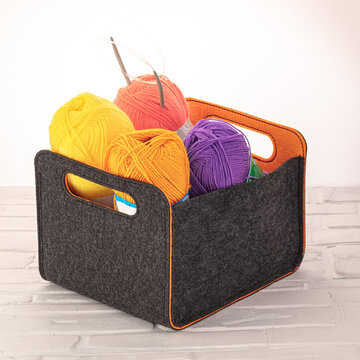 Crafting supplies are neatly arranged in a felt storage basket. Bright yellow, purple, and orange yarn balls fill the basket. Knitting needles are visible, ready for creative projects.