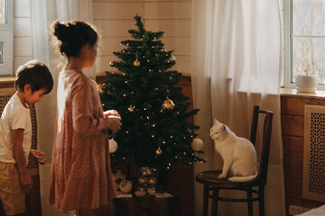 A boy and a girl are going to decorate a Christmas tree, a white cat is sitting on a chair and watching it