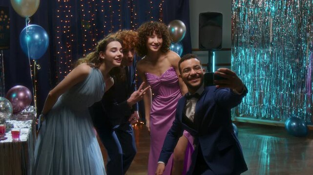 Group of cheerful teenage friends in formal suits and evening dresses taking selfies together during high school prom night party