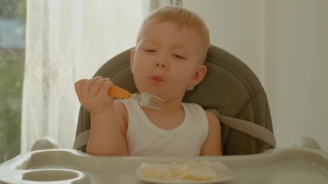 In a bright room, a little boy sits at his baby table eating fruit pieces with a fork. His curious expression shows delight as he enjoys his healthy snack.