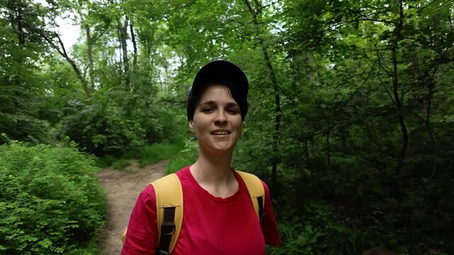 A young woman blogger walks through a green forest, filming herself with a camera. She wears a red shirt, yellow backpack and cap, enjoying outdoor adventure.