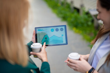 Two businesswomen holding coffee cups and reviewing tablet with business charts outdoors in modern urban environment  
