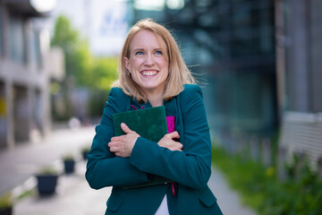 Fototapeta premium Happy businesswoman in green blazer smiling and hugging notebook outdoors in modern urban environment 