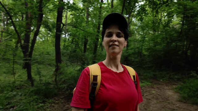 A young woman blogger walks through a green forest, filming herself with a camera. She wears a red shirt, yellow backpack and cap, enjoying outdoor adventure.