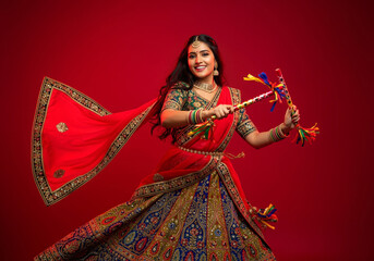 A woman garba dances in traditional outfit gracefully against a red background. She holds colorful dandiya sticks in each hand.
