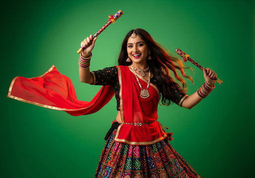 A woman garba dances in traditional outfit gracefully against a green background. She holds colorful dandiya sticks in each hand.