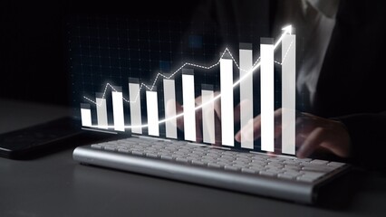 A person uses a keyboard in a dark environment while a digital graph representing business growth hovers above, illustrating technology role in data analysis. Copula