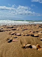 Colorful pebbles scattered across the sandy beach create a beautiful contrast against the incoming ocean waves, offering a tranquil and scenic view of the coastline on a sunny day