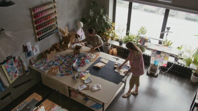 Wide top view of two Caucasian female tailors laying out paper patterns on fabric on table, preparing for cutting, while working on new design in atelier, next to patchwork garments and cloth swatches