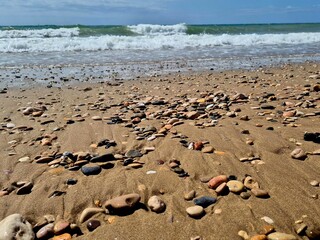 Colorful pebbles scattered across the sandy beach create a beautiful contrast against the incoming ocean waves, offering a tranquil and scenic view of the coastline on a sunny day