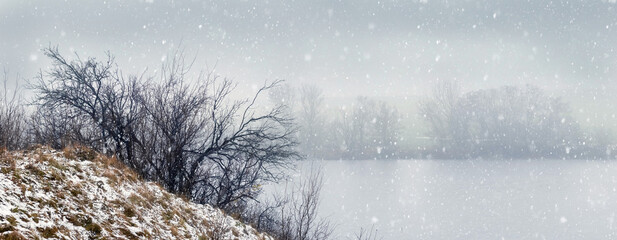 Winter landscape with falling snow over lake and shore covered with bushes misty and quiet...