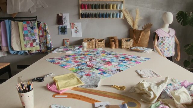 Full no people shot of interior of sewing workshop, with wall display of multicolored thread on bobbins, scraps of cotton fabric on large cutting table. Ideal for atelier and tailor shop ads 