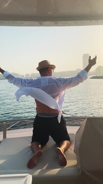 Handsome man with hat enjoying sunset sail on luxury yacht in Dubai Marina with hands wide open up in the air, vertical video
