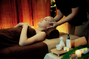 Caucasian woman enjoying relaxing anti-stress head massage and pampering facial beauty skin recreation leisure in warm candle lighting ambient salon spa in luxury resort or hotel. Quiescent