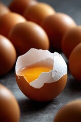 Close-up of cracked egg with yolk surrounded by whole brown eggs on dark surface
