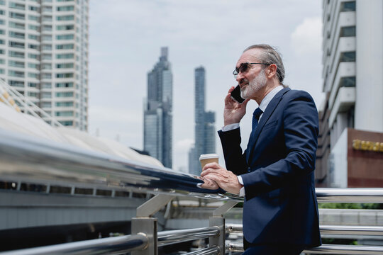 Confident senior businessman in suit holding coffee and using smartphone for video call in modern city, symbolizing leadership, communication, technology, and professional lifestyle.
