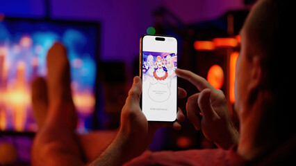 Reading colored maid girls digital manhwa comic webtoon on smartphone with RGB PC in background, over shoulder view