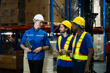 Three warehouse workers of different ethnic backgrounds communicating while holding tablet and clipboard during warehouse operation near stacked goods and storage shelves