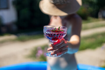 Woman in a hat enjoys a colorful cocktail while relaxing by an outdoor pool on a sunny summer day in a garden setting