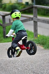 Toddler boy in green shirt and neon helmet rides red balance bike uphill on asphalt path. Active...
