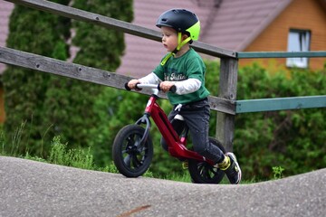 Obraz premium Little boy with neon helmet and green t-shirt riding red balance bike on track. Focused toddler learning cycling skills and enjoying outdoor activity in park environment.