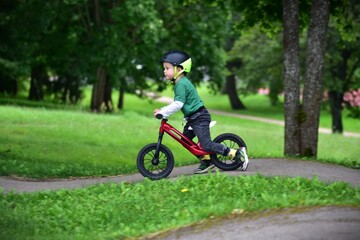 Smiling toddler riding a red balance bike on a park path, wearing a helmet for safety. Active child enjoying outdoor fun, learning to balance and cycle independently in a green natural environment.