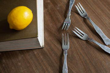 Four small forks and a lemon placed beside a book on a wooden table during an afternoon light