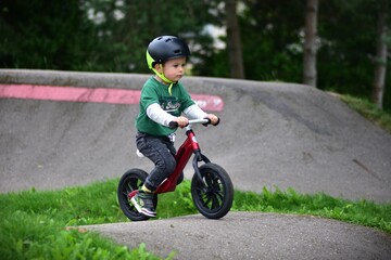 Small boy rides red balance bike on pump track in park. Child wearing helmet practices cycling, balance, and coordination while enjoying outdoor adventure and healthy activity.