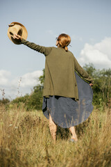 Woman enjoying a sunny day in a field while holding a straw hat and wearing a casual outfit suitable for warm weather