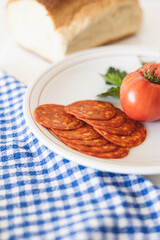 Sliced chorizo and fresh tomato on a white plate with a checkered cloth and loaf of bread in the background during a sunny afternoon