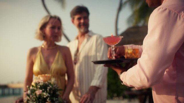 A waiter carries a tray of colorful cocktails toward a mature couple at a luxurious tropical beach resort. This golden hour scene highlights hospitality, vacation indulgence, and upscale travel.