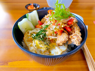 Close-up of a delicious Asian-style rice bowl with crispy fried chicken, sunny side up egg, spicy chili garnish, and fresh cucumber slices. Served in a black ceramic bowl on a wooden table.