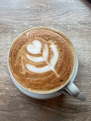 Close-up of a latte with intricate coffee art resembling a flower or leaf on top, served on a wooden café table, capturing a cozy and artistic coffee experience