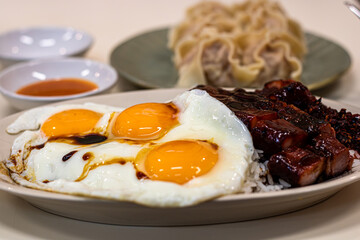 Close-up of a plate with fried sunny-side-up eggs and runny yolks served alongside glazed char siu (roasted pork) on steamed white rice, with steamed dumplings and dipping sauce in the background. War