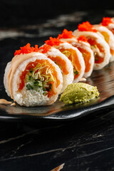Close-up of sushi rolls with salmon, cream cheese, cucumber, lettuce, and white rice. Topped with red fish roe and served with wasabi on a black plate. Elegant Japanese cuisine and restaurant presenta